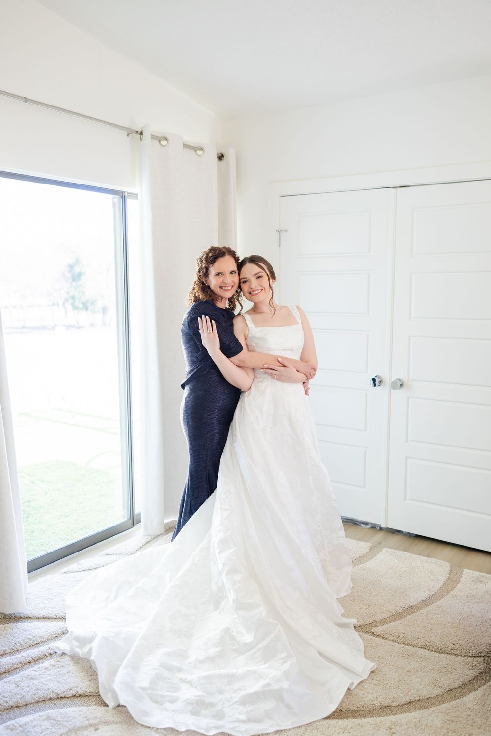 Two people embrace in a bright room with white walls and window, one wearing a long flowing white wedding gown with train.