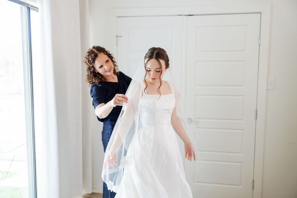 Wedding day preparation photo showing a bride in a white dress and veil while being assisted with final touches.