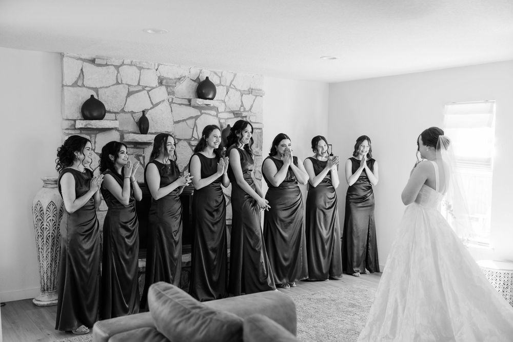 A bridal party in matching charcoal dresses stands in a row watching a bride in a white gown during wedding preparations.