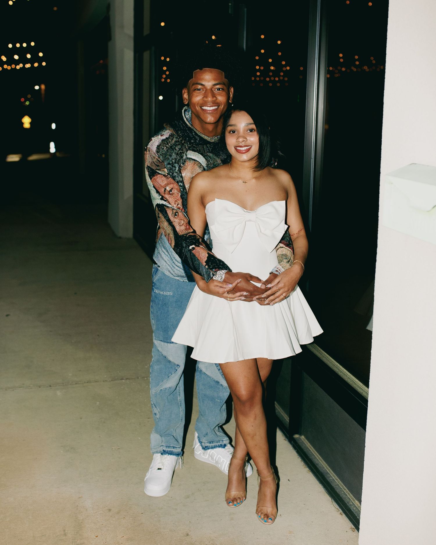 A couple poses together at night, with the person in a white strapless dress and another in jeans and a patterned shirt.