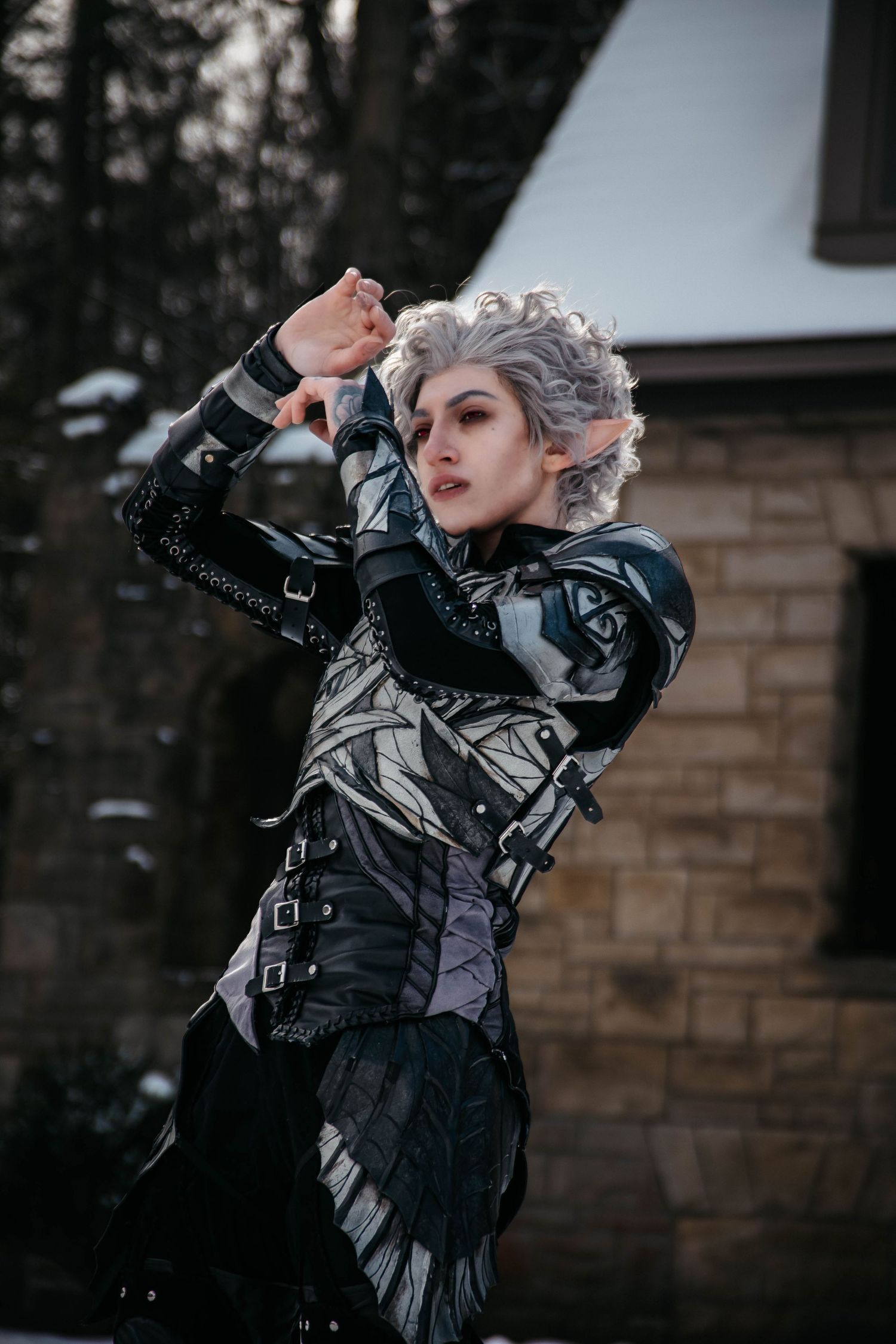 A person in a gothic black leather costume with silver hair poses dramatically against a dark winter backdrop.