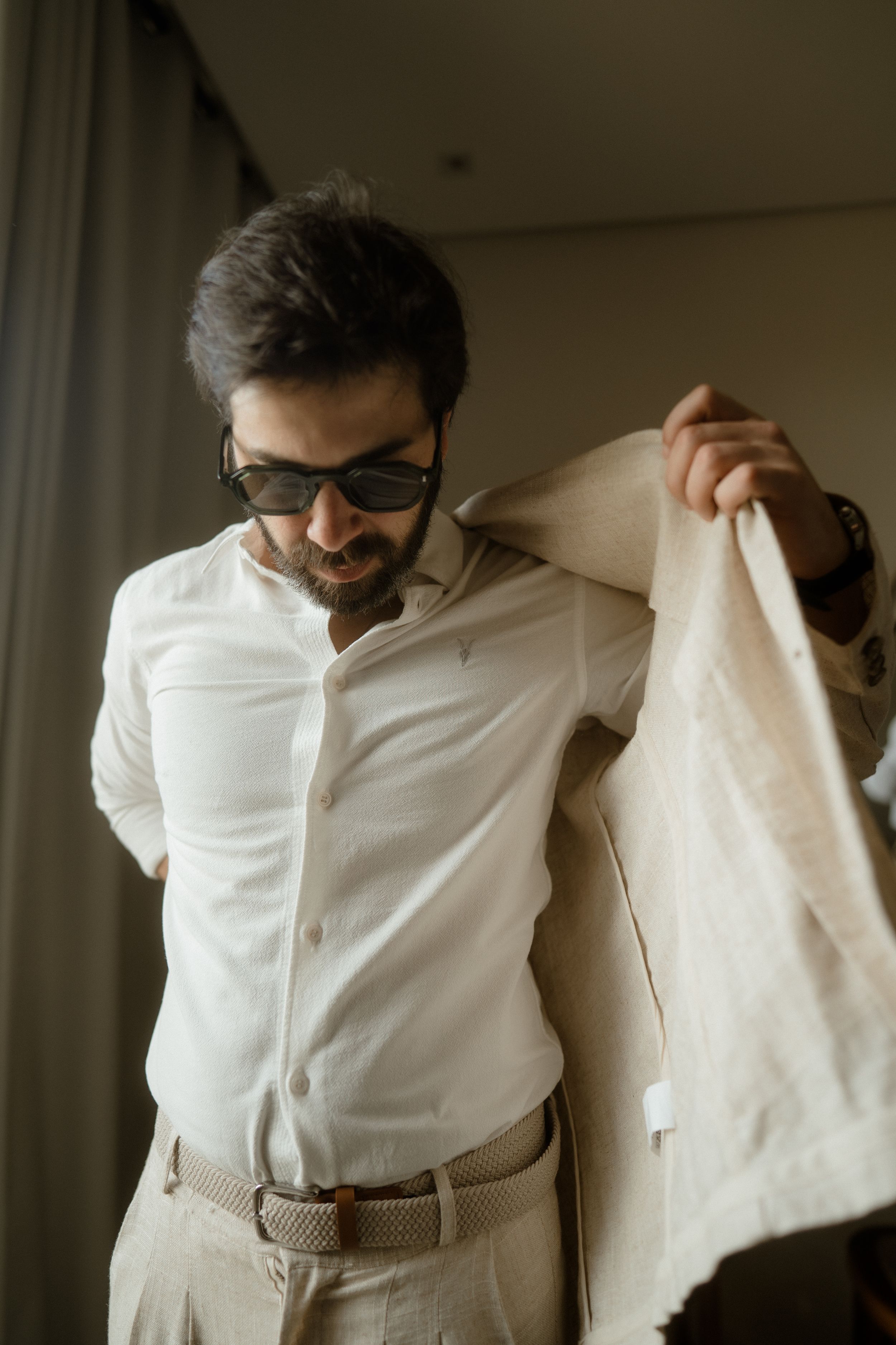 A person in a white button-up shirt and sunglasses puts on a light colored jacket in an indoor setting.