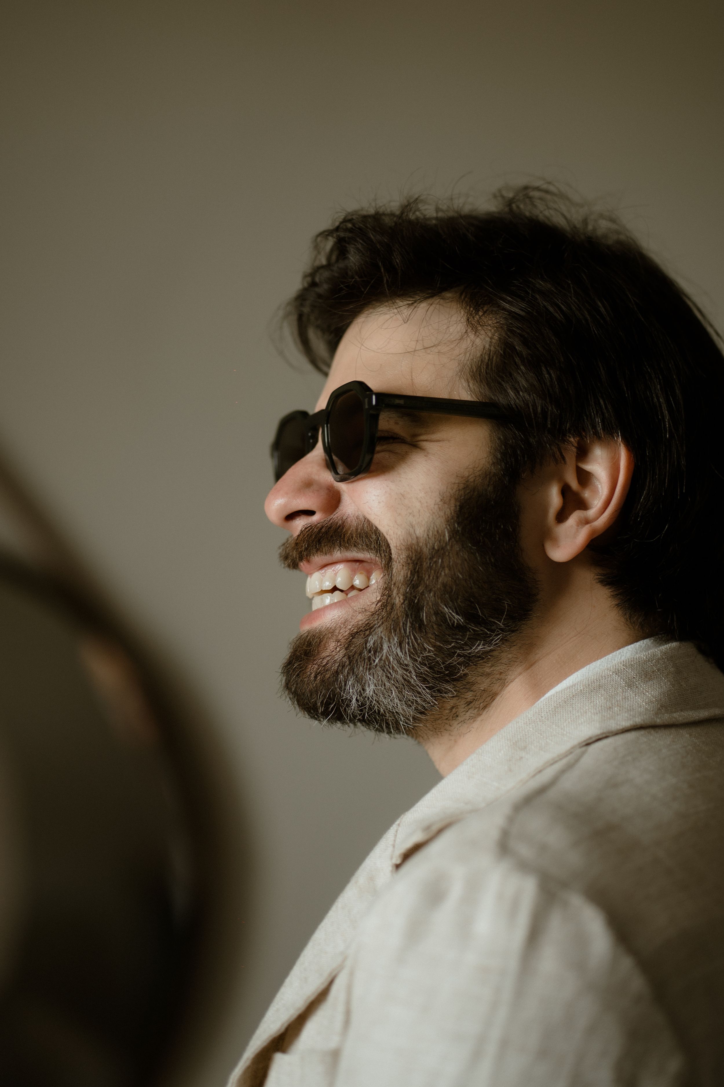 A bearded person in sunglasses and light clothing smiles warmly in a candid profile shot.