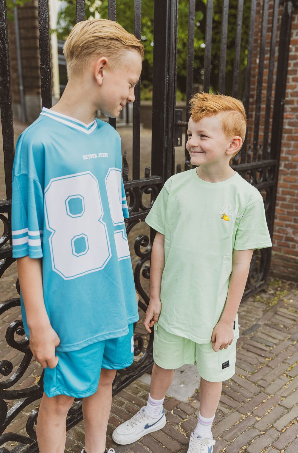 Two young people in casual summer outfits standing on a brick path and smiling at each other near an ornate iron gate.