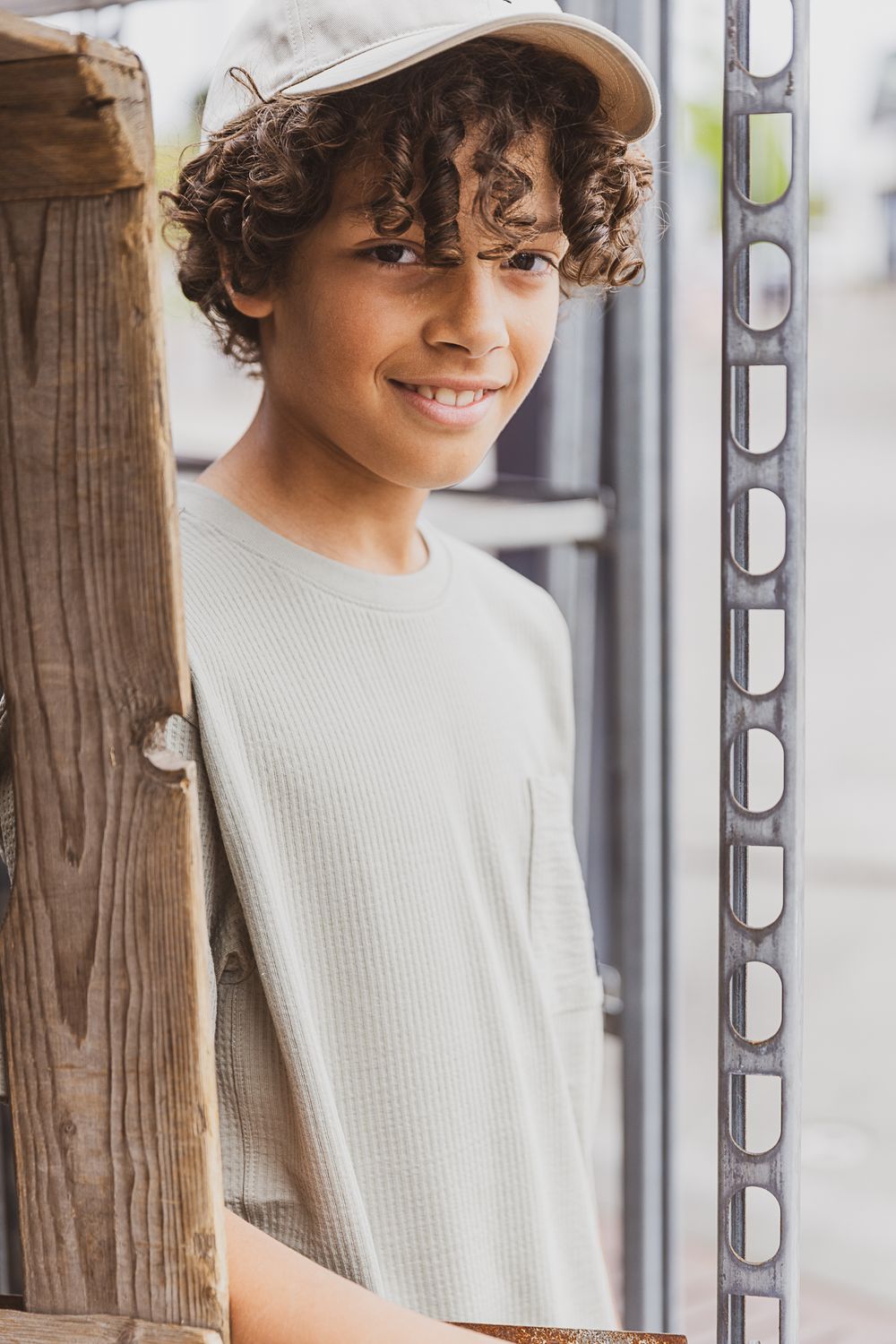 Young person in a beige sweater smiling while standing by a wooden post and window.