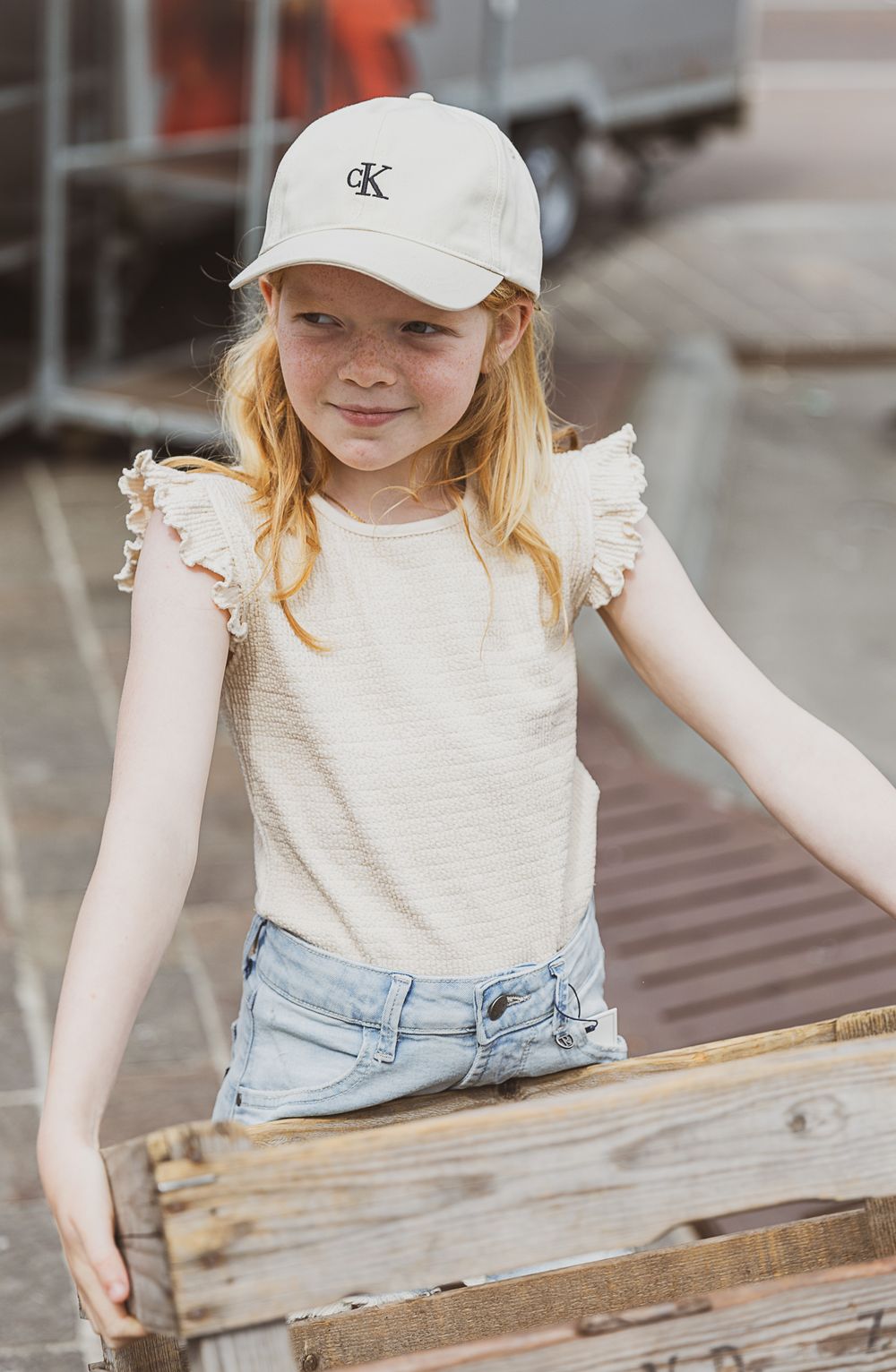 Young person wearing a white baseball cap and ruffle sleeve top holds a rustic wooden crate outdoors.