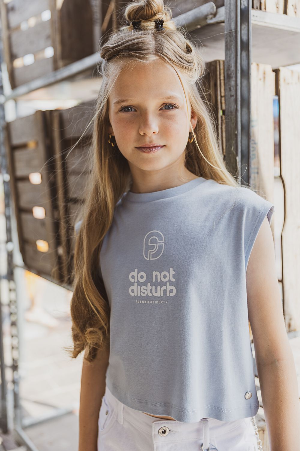 Young person with long blonde hair wearing a light gray sleeveless t-shirt with 'do not disturb' text in natural lighting.