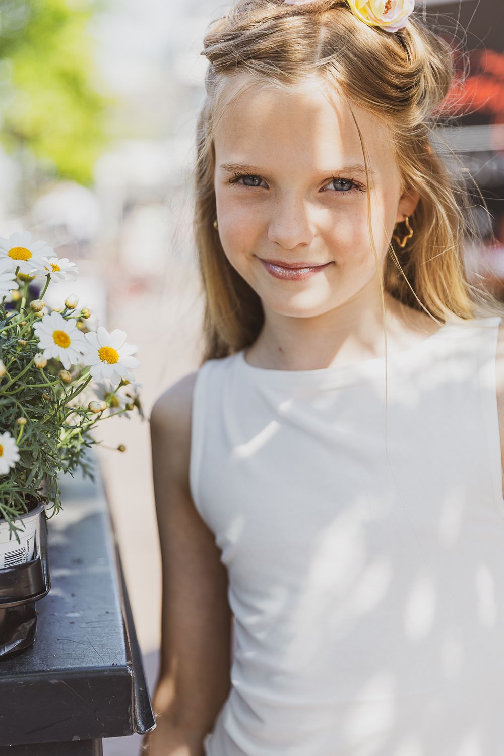 Young person in white sleeveless dress standing next to potted daisies in a sunny outdoor setting.