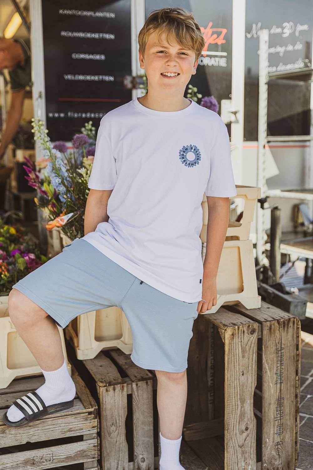 A young figure in a white t-shirt and light blue shorts sits casually on wooden crates near a storefront with flowers.