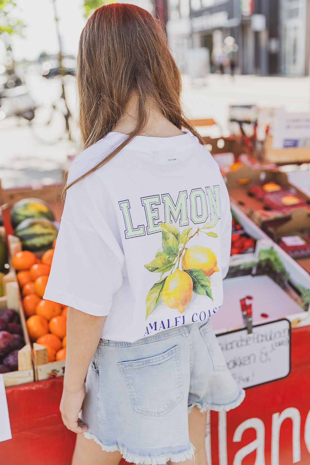 Woman in white t-shirt with lemon graphic shops at outdoor produce market with colorful fruits and vegetables on display.