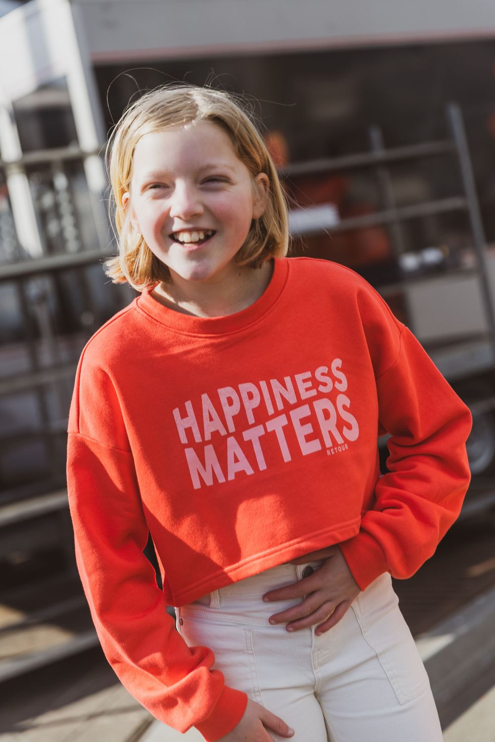 Someone wearing a bright orange cropped sweatshirt with 'HAPPINESS MATTERS' text and white pants poses outdoors.