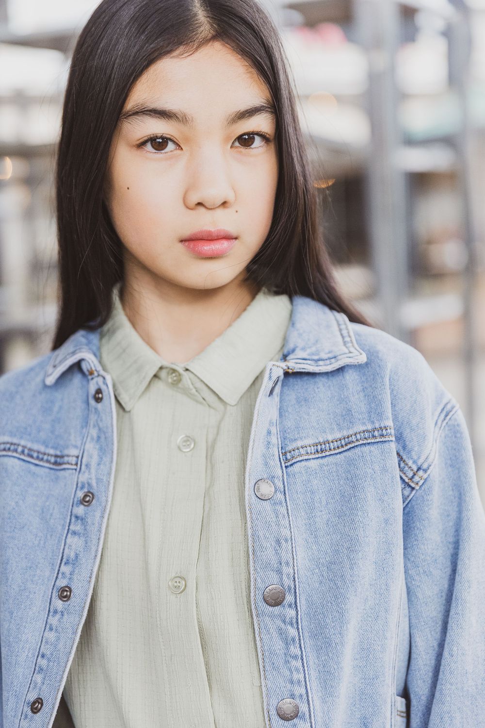 Portrait of person in denim jacket and light button-up shirt against blurred urban background.