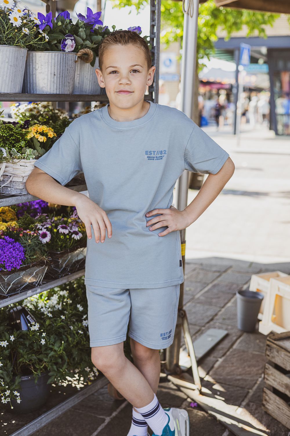 Child wearing light blue matching shorts and t-shirt outfit poses next to colorful flower display at outdoor market.