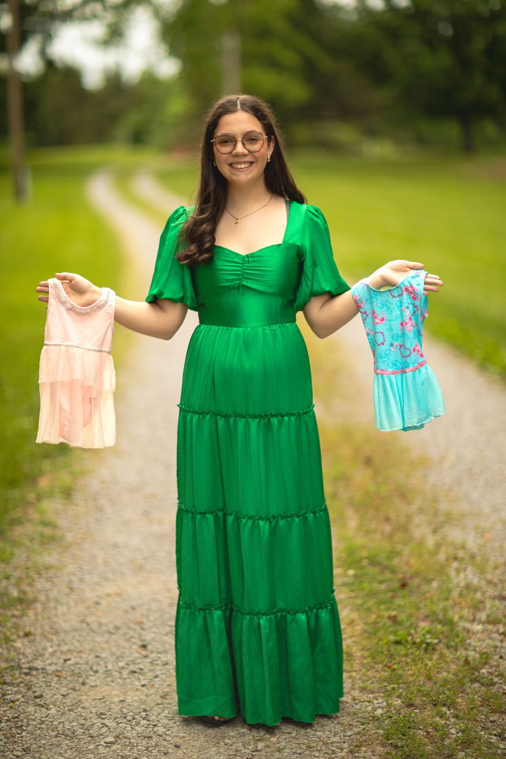 Graduate in emerald green tiered dress holding up two small dress outfits on a gravel path.