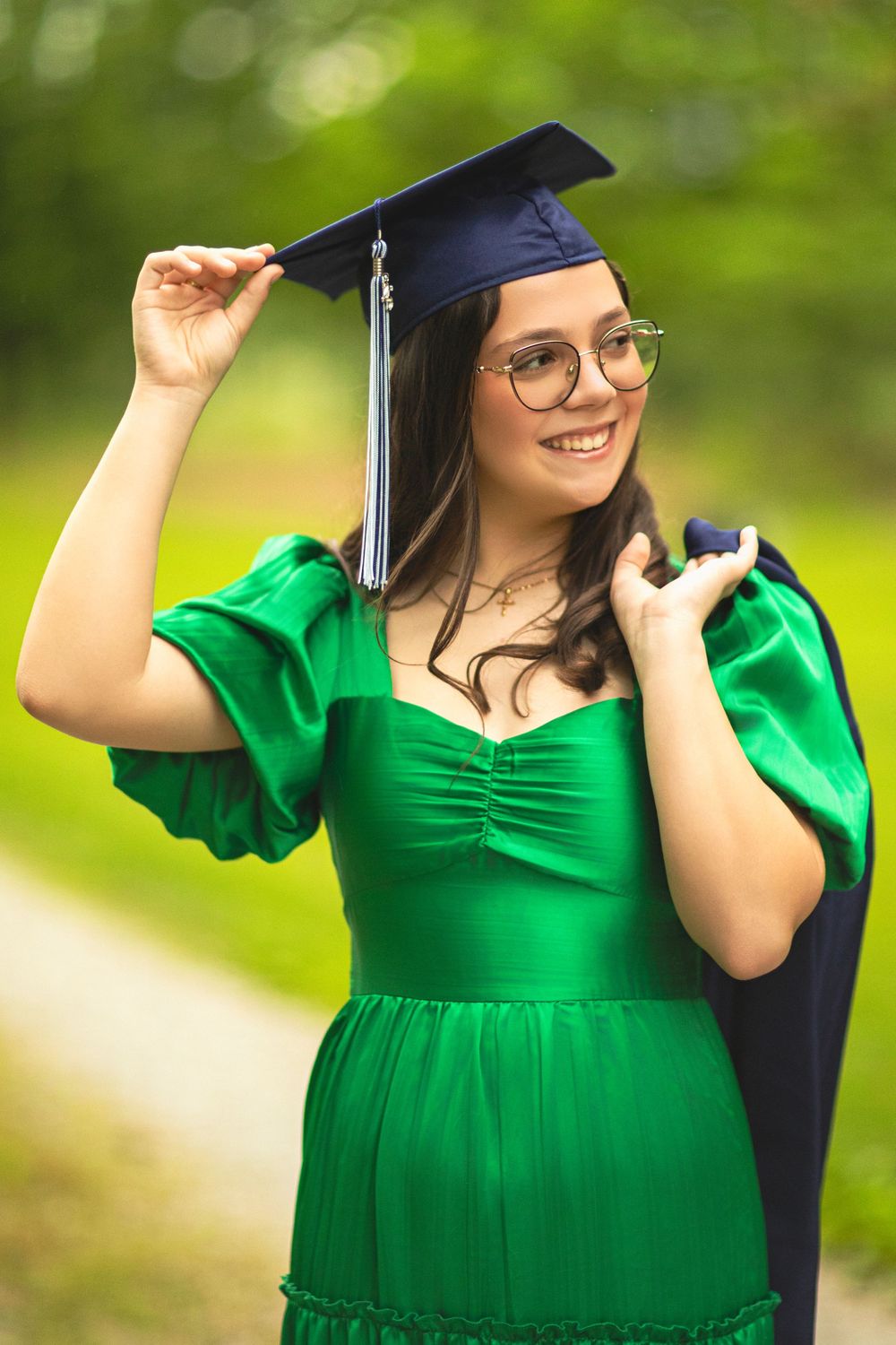 Graduate in emerald dress and cap smiling while adjusting mortarboard outdoors.