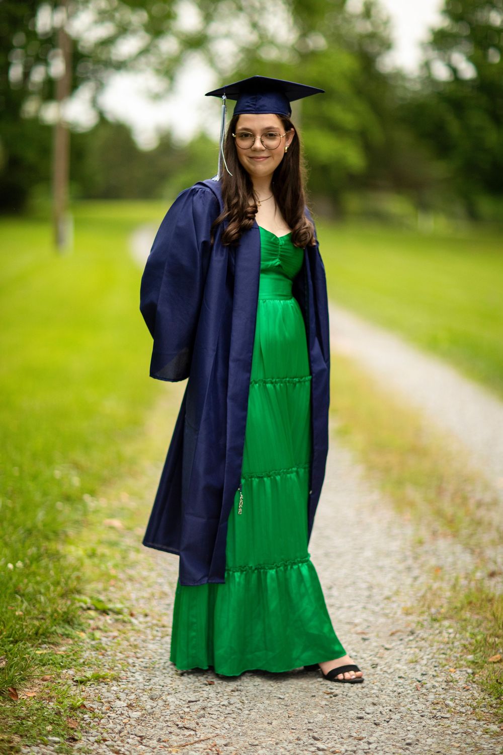 Graduate wearing navy graduation gown over emerald tiered dress stands on outdoor path.