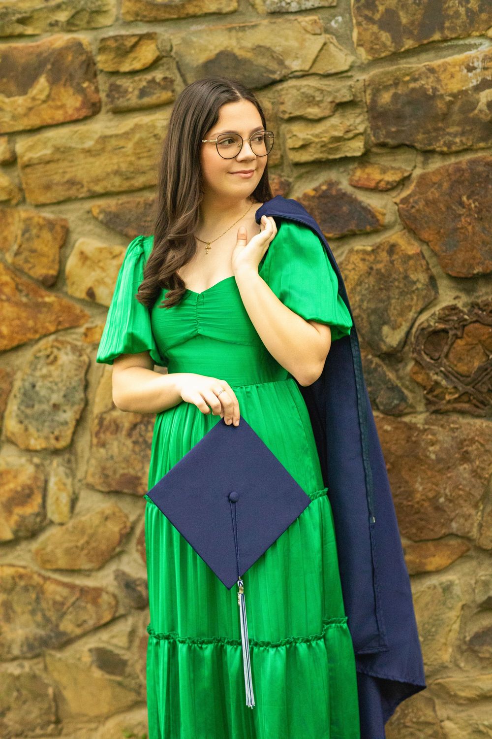 Graduate in flowing green dress poses with diploma against stone wall backdrop.