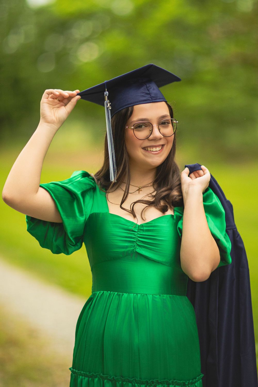 Graduate adjusting mortarboard cap while wearing emerald green dress and glasses.