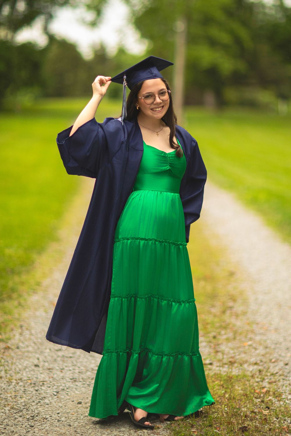 Graduate in emerald dress and navy cap and gown stands on gravel path.