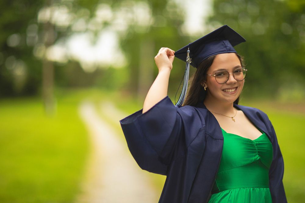 Graduate in emerald dress and navy graduation gown poses outdoors.