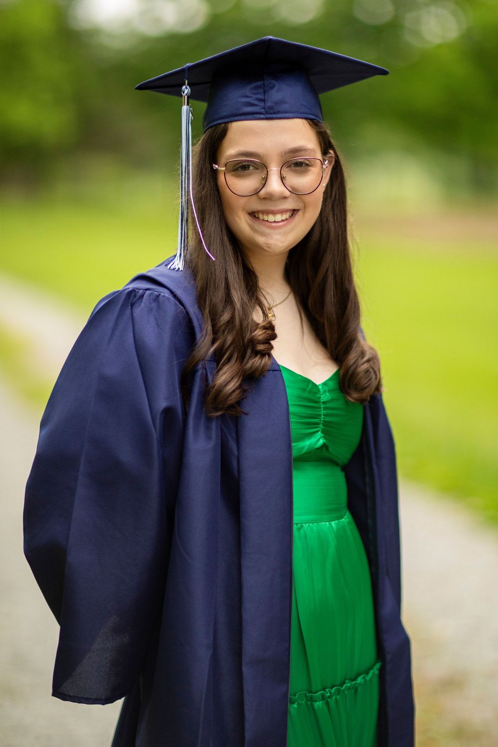 Graduate in navy cap and gown with emerald dress poses on outdoor path.