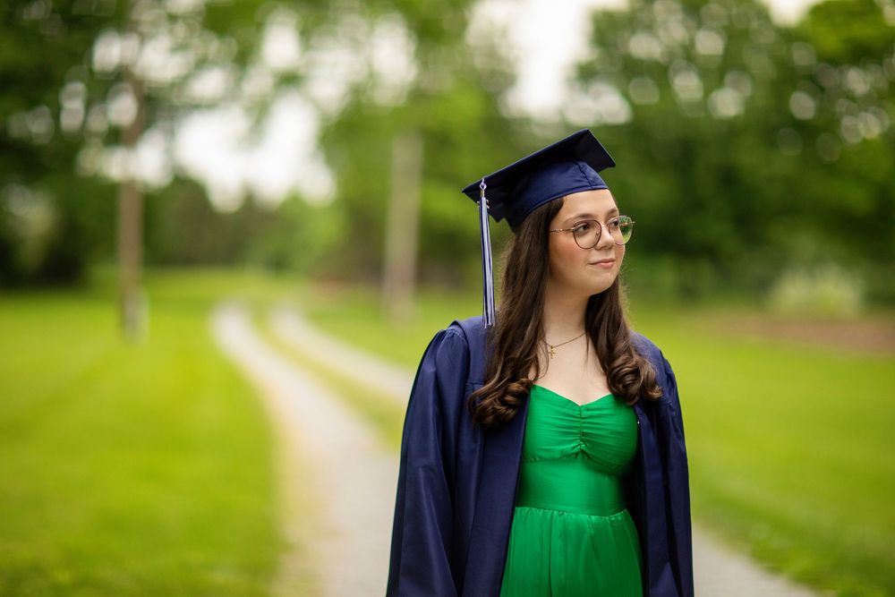 Graduate in navy cap and gown and bright green dress poses in a park setting.