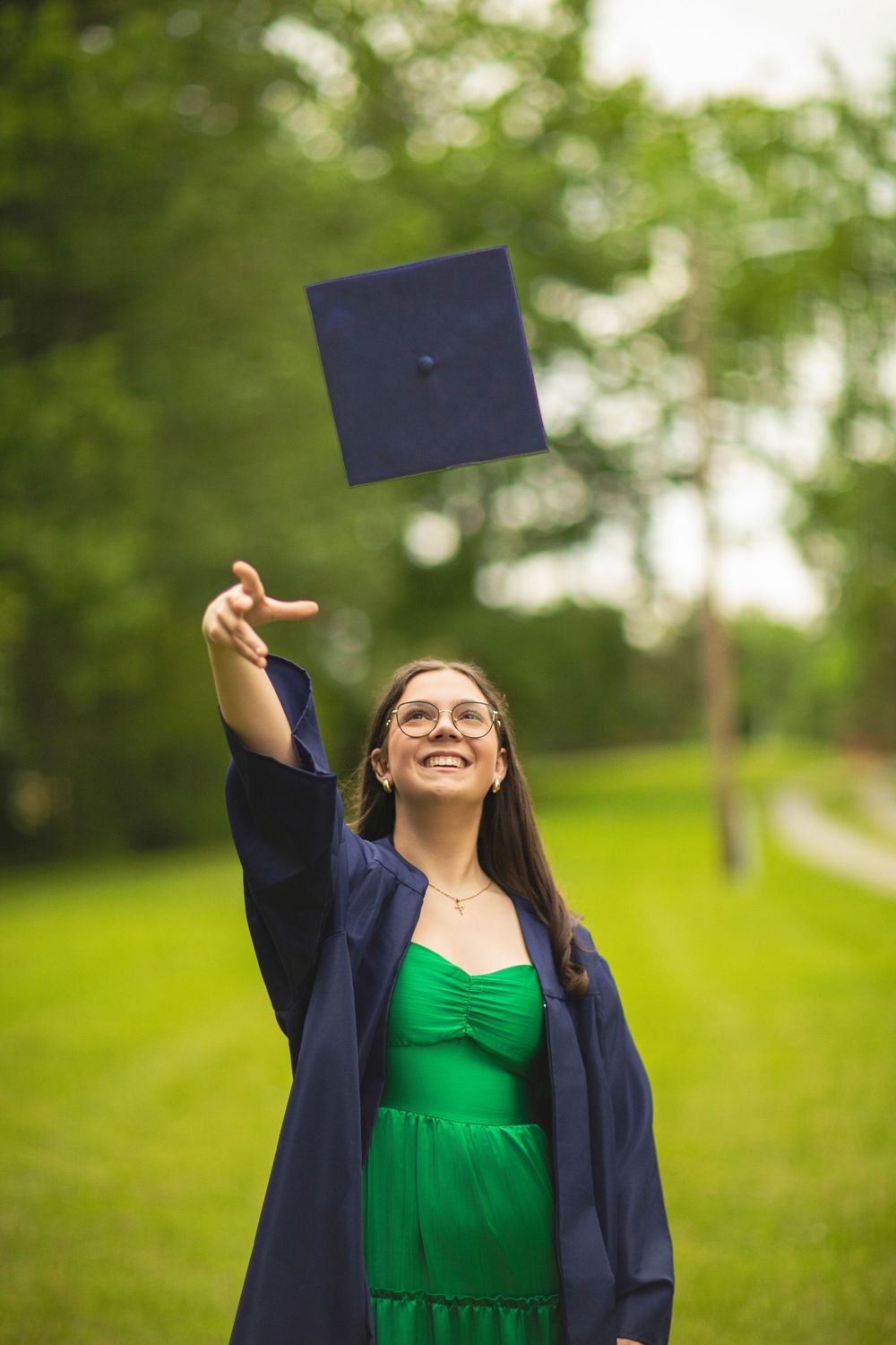 Graduate tossing graduation cap in air while wearing navy gown and green dress outdoors.