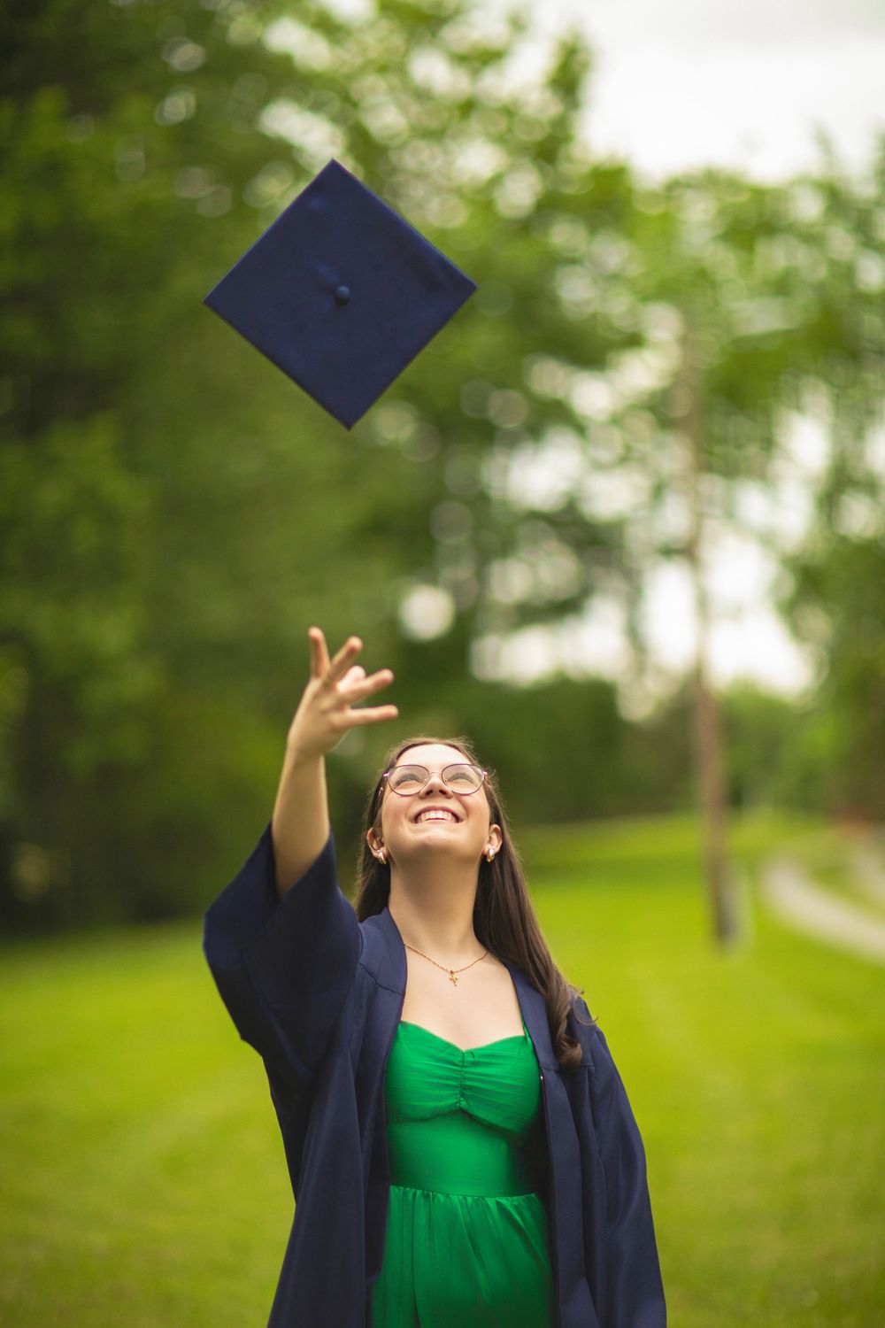 Graduate celebrating by throwing cap in air against green outdoor backdrop.