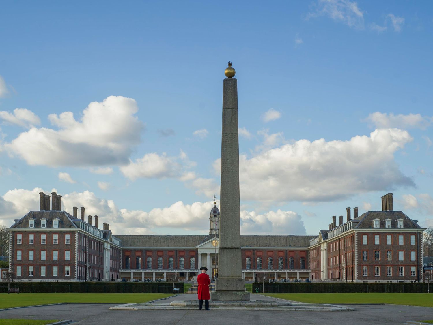 A tall white obelisk monument stands between two symmetrical brick buildings at Royal Hospital Chelsea on a sunny day.