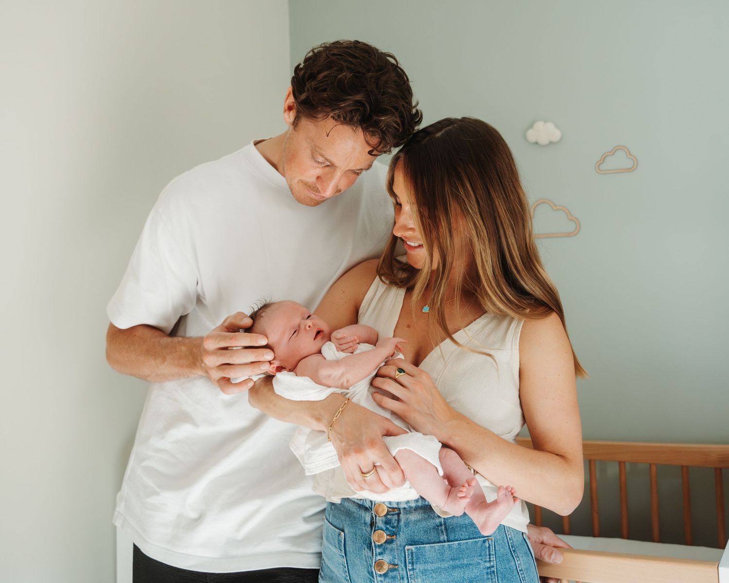 New parents lovingly cuddle their newborn baby together in a nursery with soft cloud wall decorations.