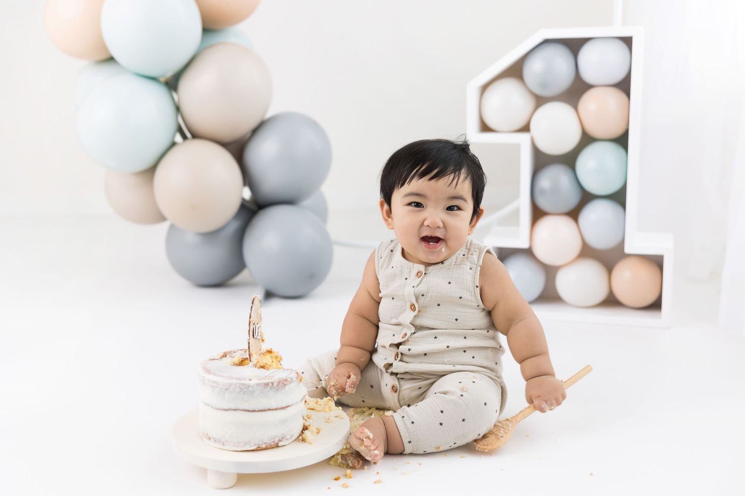 Baby in cream-colored outfit sits on white floor with pastel balloon decor and birthday cake for first birthday celebration.