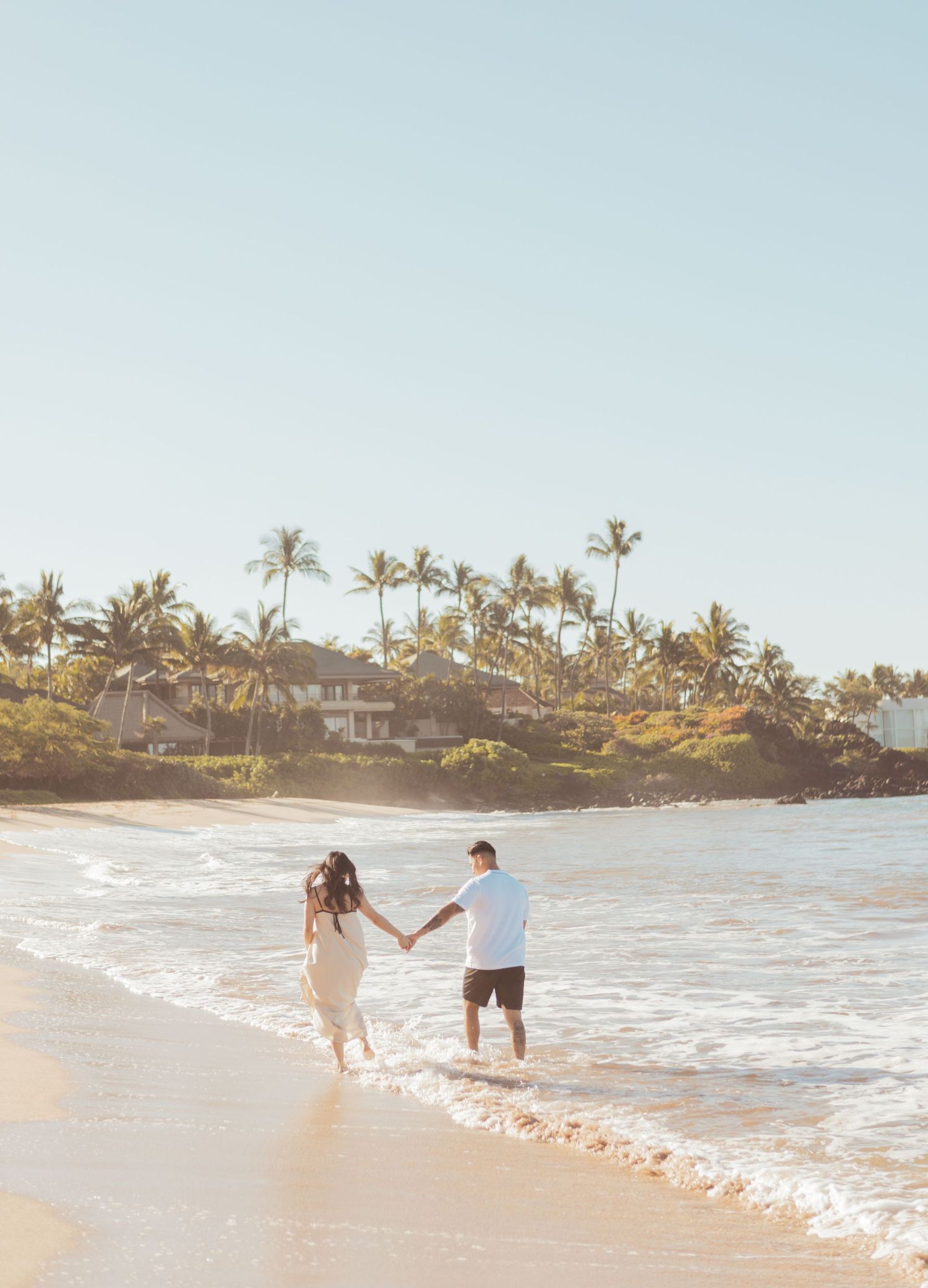 Two figures hold hands while walking along a scenic Hawaiian beach with palm trees and oceanfront homes in the background.