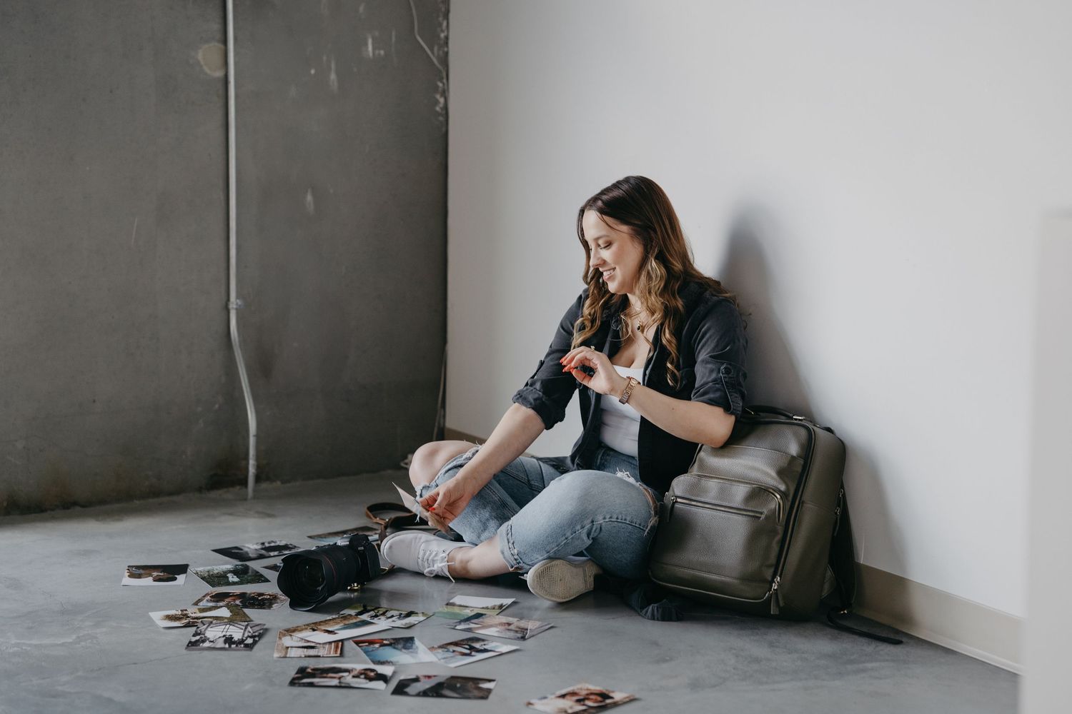 Person sitting on floor against wall sorting through scattered photographs while holding smartphone.