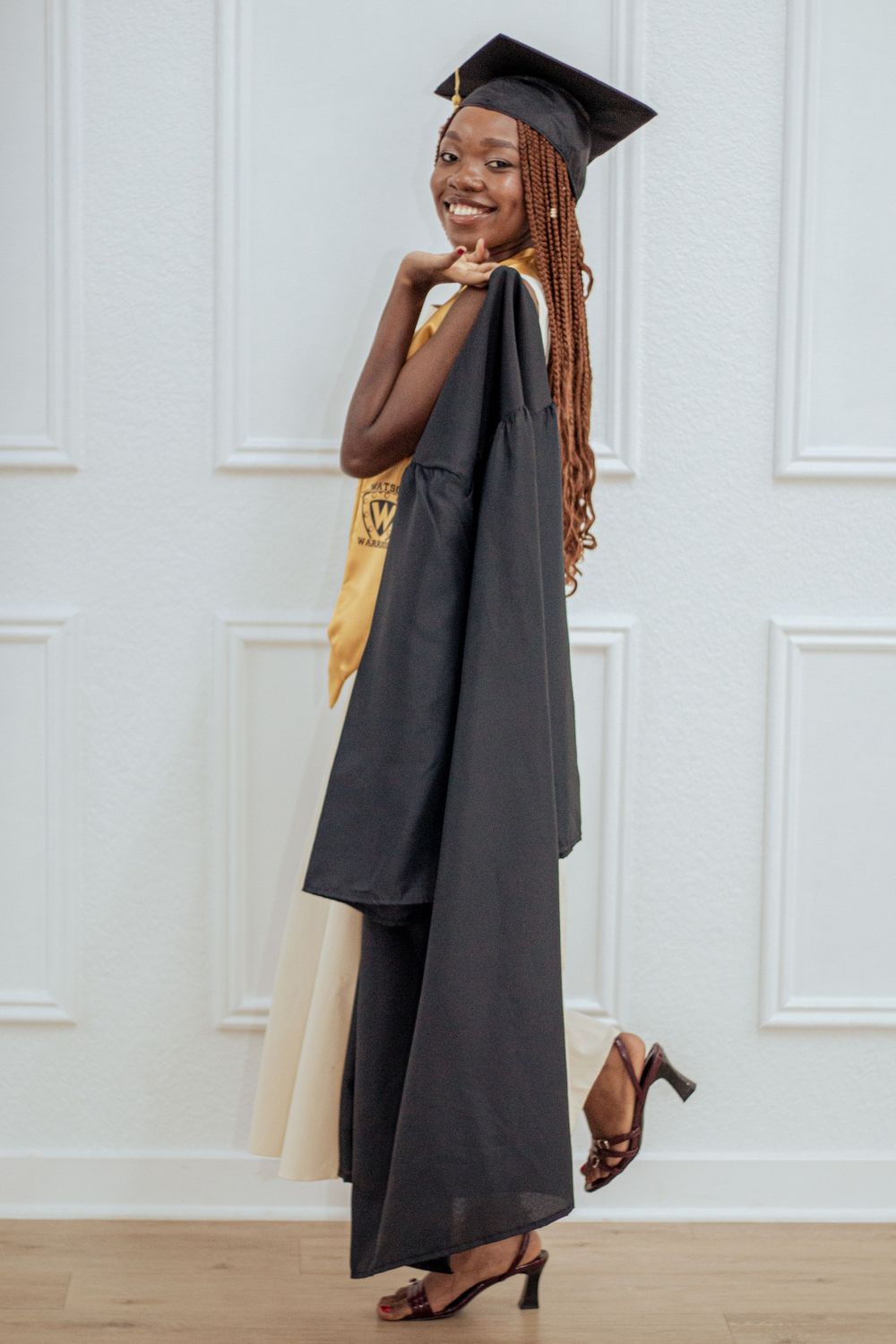 Graduate in black cap and gown with gold stole poses elegantly against white wall during commencement celebration.