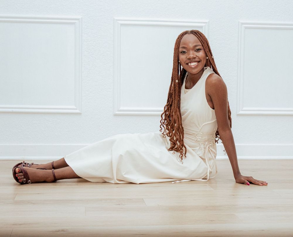 A woman in an elegant white dress reclines on a hardwood floor against a white paneled wall while smiling warmly.
