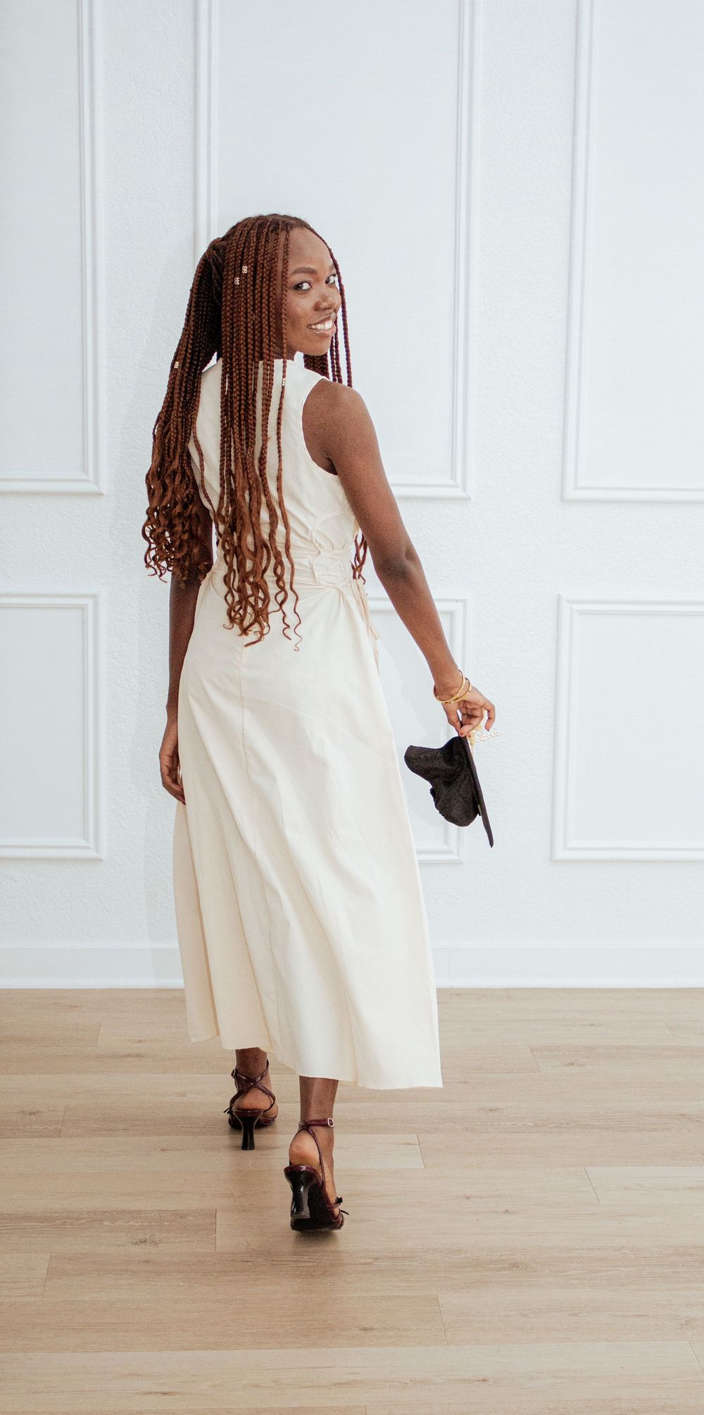 A person in an elegant cream-colored sleeveless dress and heels poses against a white wall with hardwood floors.