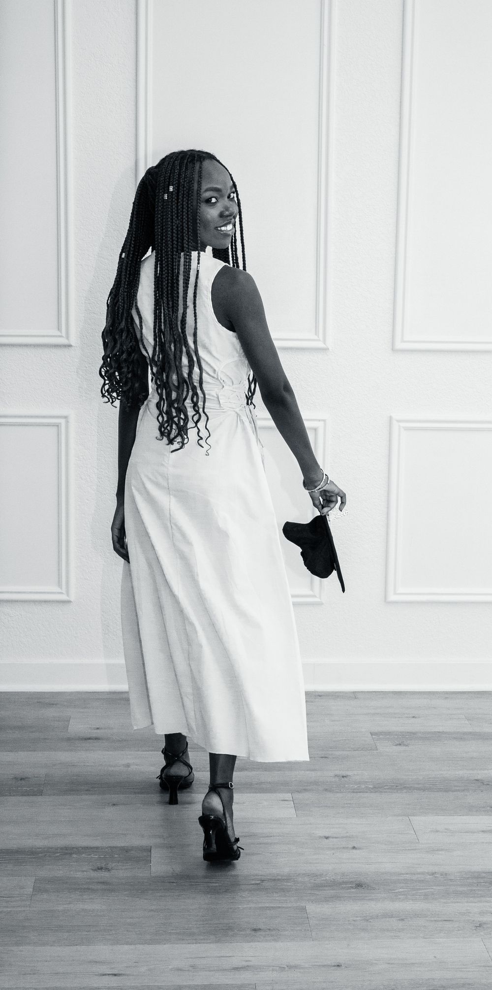 A black and white photo shows a figure in a flowing white dress and heels posing against a paneled wall.