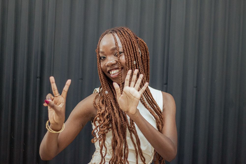 Woman with long braids smiling and making peace sign gesture against dark grey wall.