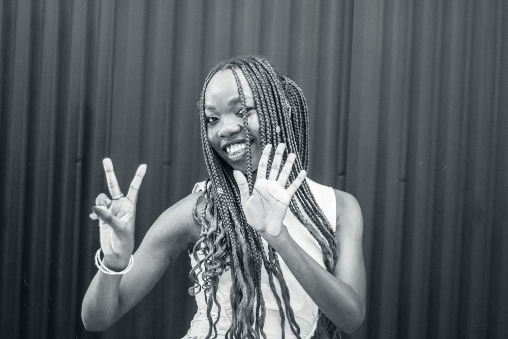 A portrait of someone with long braids smiling and showing a peace sign in front of a corrugated wall.
