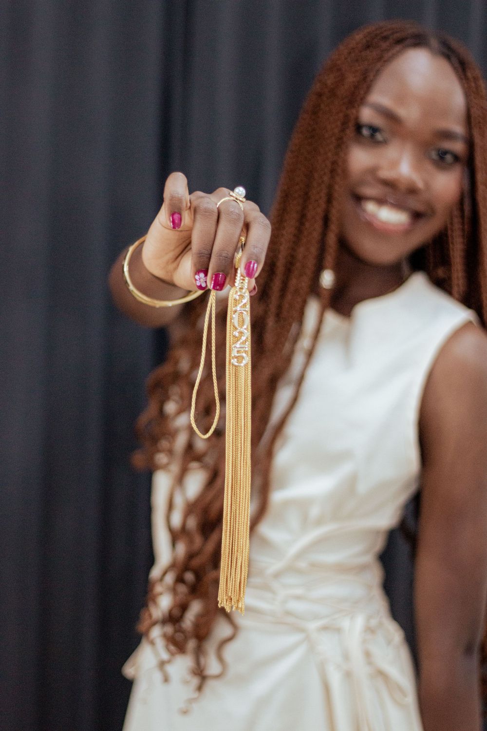A graduate holding up a gold graduation tassel while wearing a white dress and displaying long braided hair.