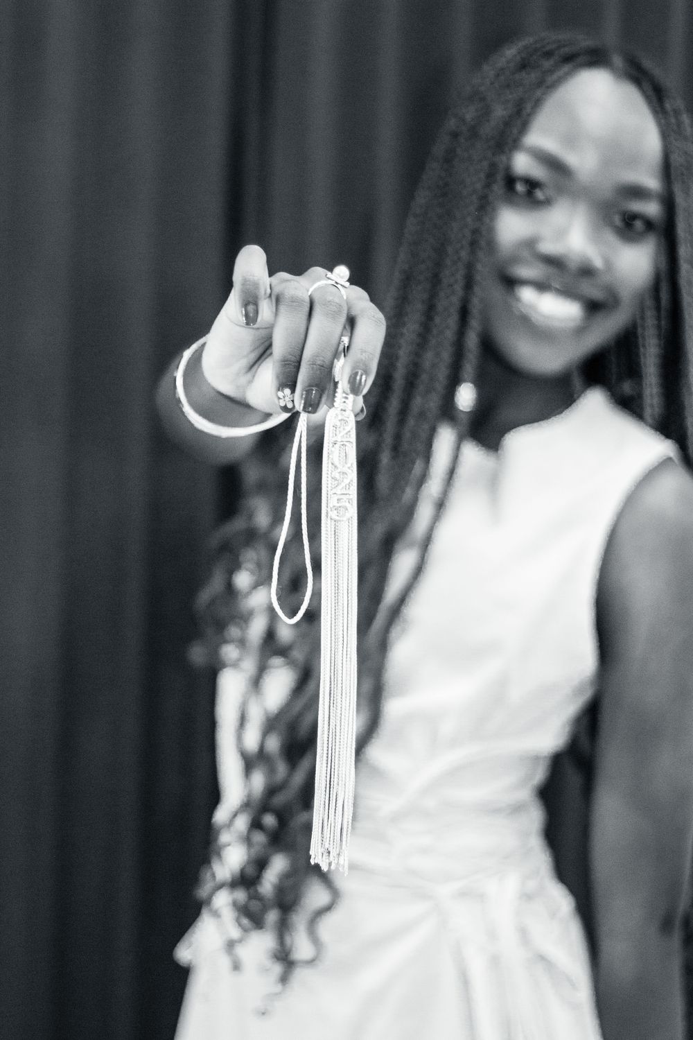 Graduate in white dress displays graduation tassel and cord with joyful expression in black and white photo.