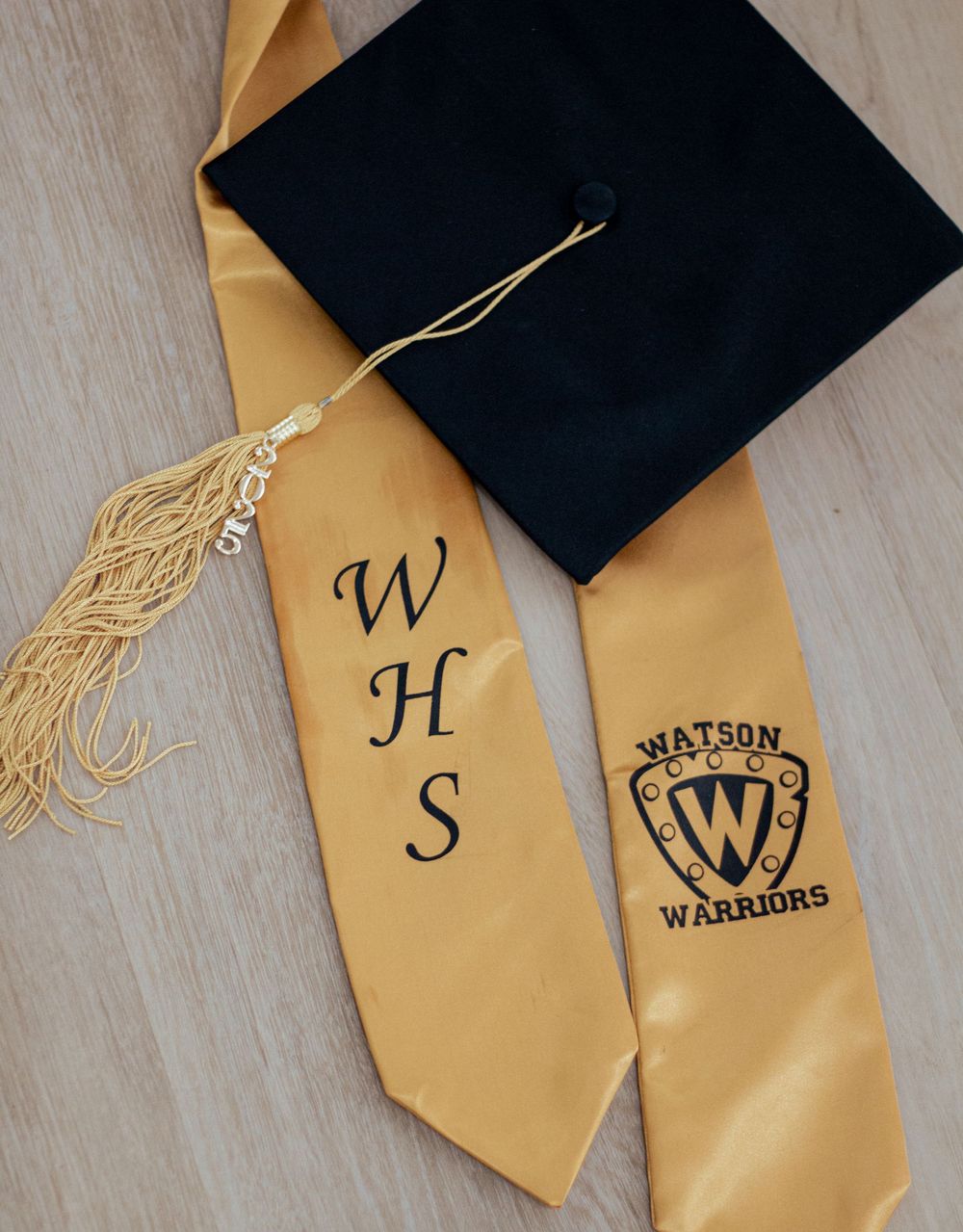 Black graduation cap with gold tassel and gold honor stole showing WHS Watson Warriors school emblem.