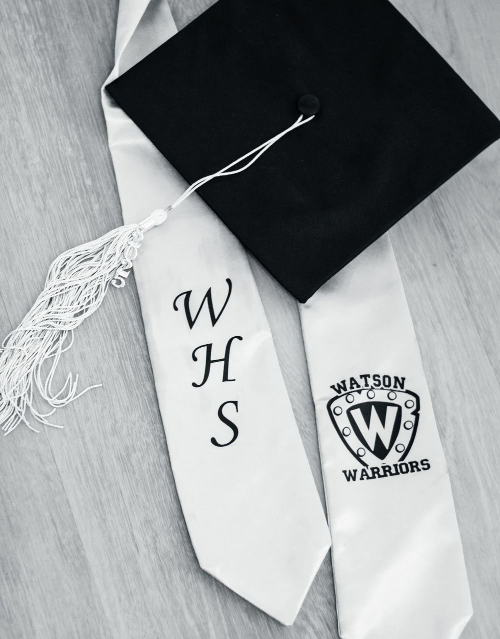 A black graduation cap with white tassel and school stole showing WHS initials and Watson Warriors logo.