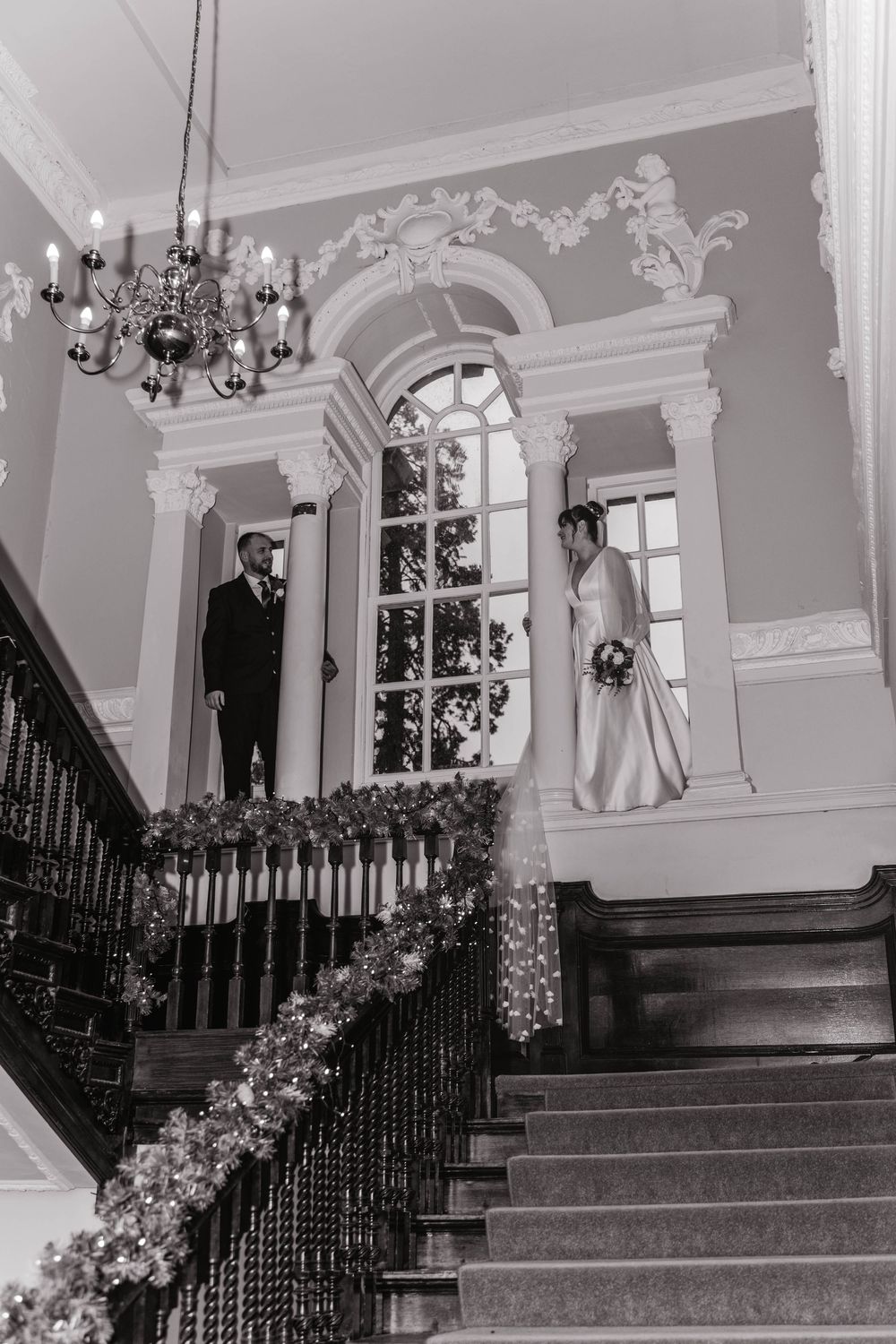 Elegant black and white wedding photo on ornate staircase with chandelier and floral decorations.