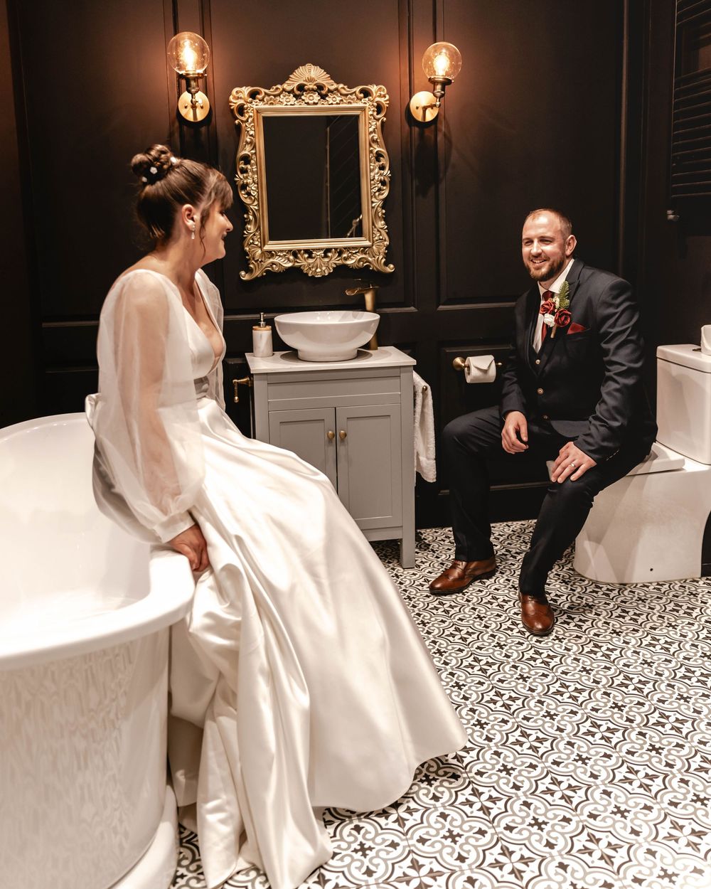 A couple shares a moment in an elegantly decorated bathroom with patterned tile floors and vintage fixtures.