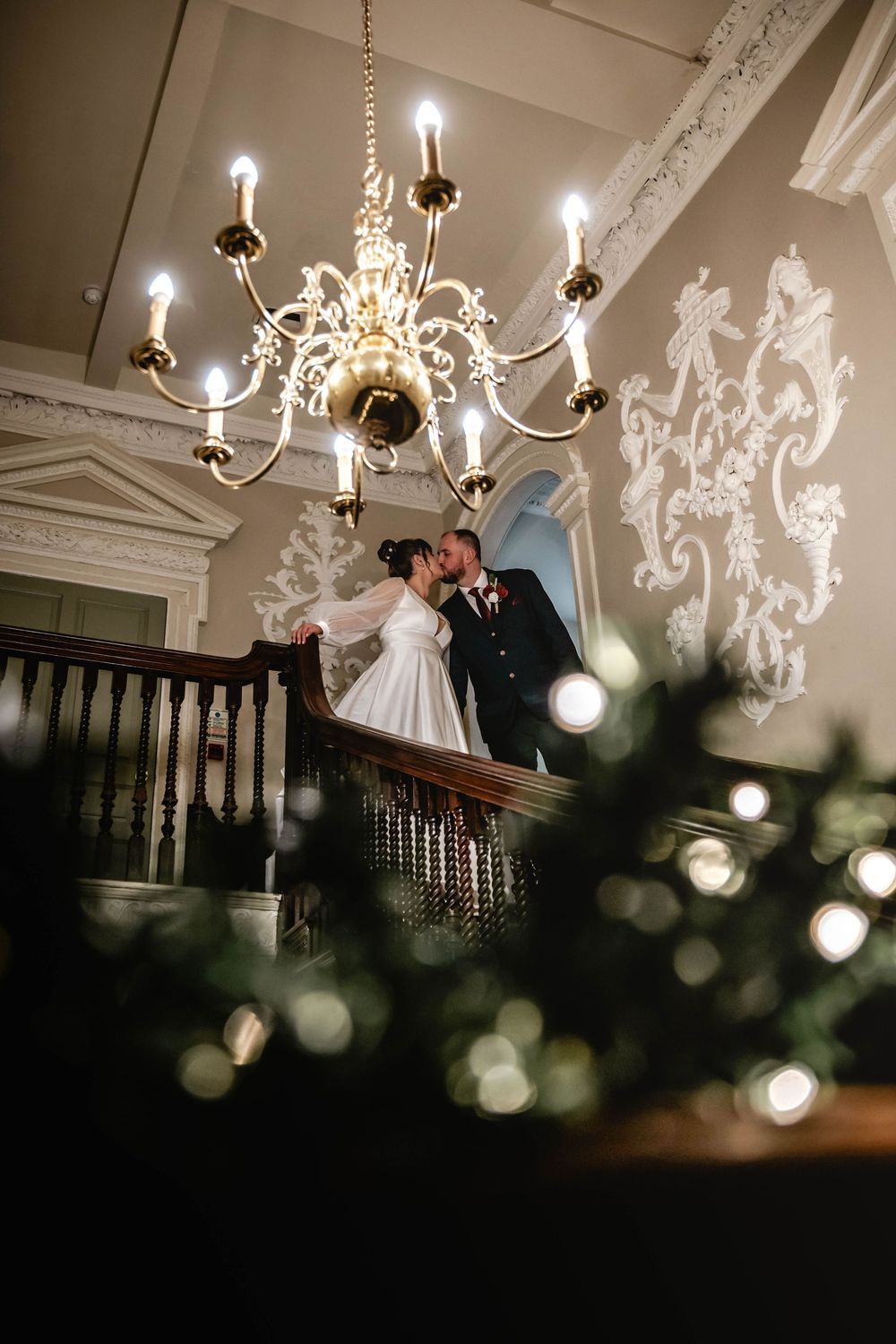Newlyweds share a romantic moment under an elegant chandelier on a grand staircase.