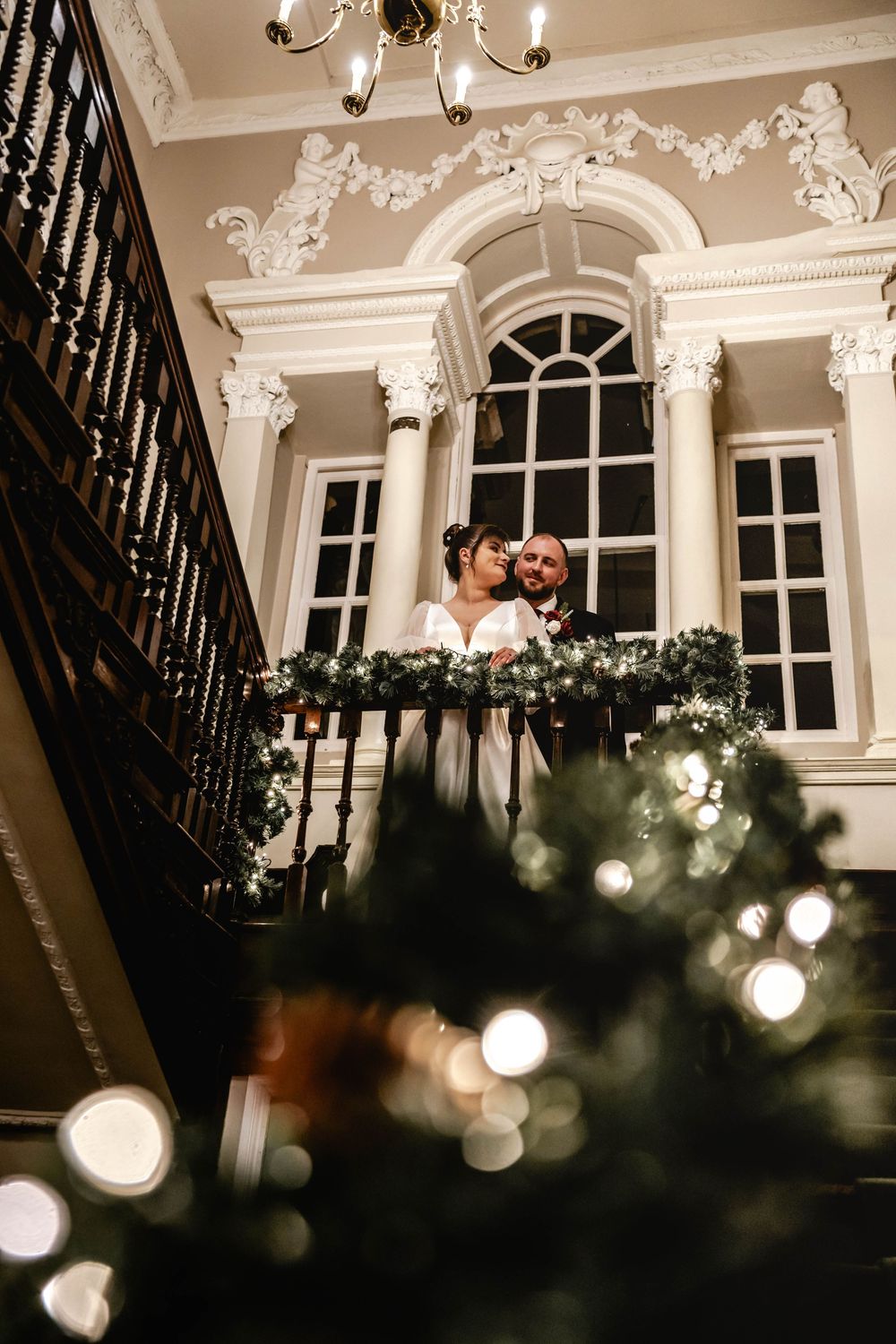 Wedding couple poses on an ornate staircase with decorative moldings and bokeh lights.