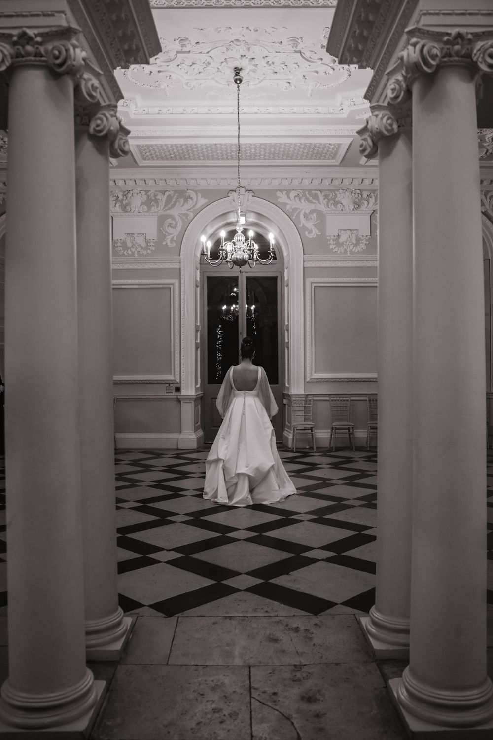 A wedding gown flows elegantly through a classical corridor with black and white checkered floors.