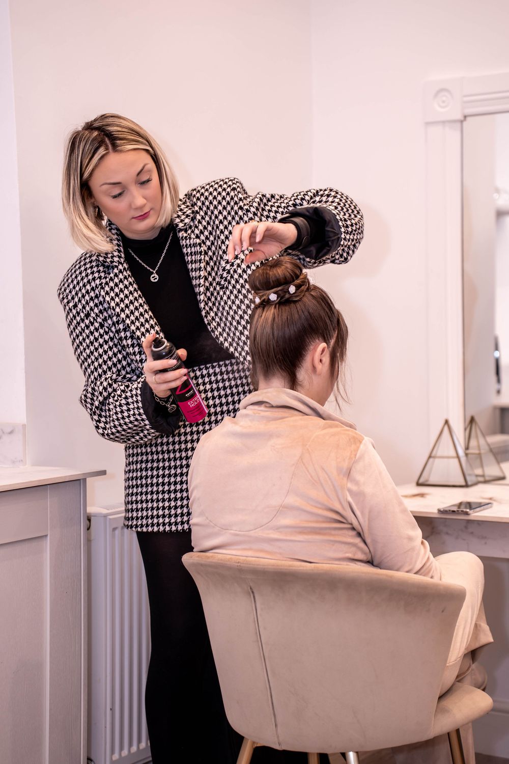 Stylist in houndstooth blazer working on client's hair in bright salon setting.