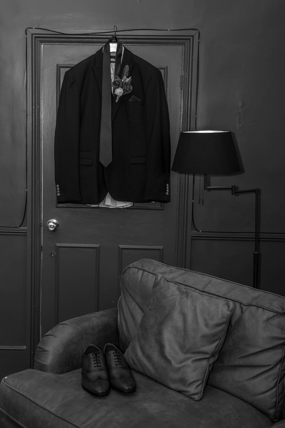 A formal suit jacket and tie hanging on a door with dress shoes placed near a couch.